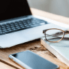 Image of a laptop, wireless phone, notebook and glasses on a desk for a blog post discussing mystery shopping as a side hustle.