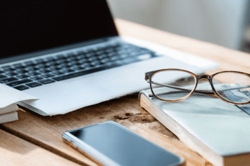 Image of a laptop, wireless phone, notebook and glasses on a desk for a blog post discussing mystery shopping as a side hustle.