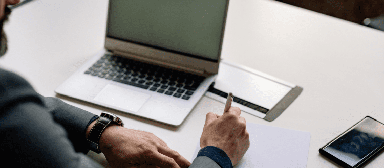 Image of a man at a desk working on his laptop and taking notes for a blog post covering mystery shopping preparation.