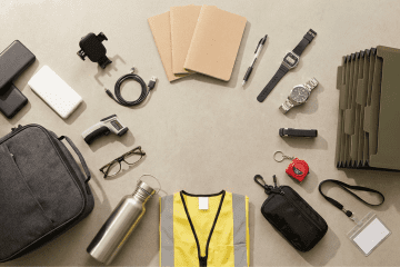 Image of a mystery shopper carry kit laid out in a flat-lay arrangement, including a portable charger, car phone mount, notebooks, pens, two Casio watches, a file folder, EDC pouch, insulated lunch bag, water bottle, safety vest, infrared thermometer, reading glasses, keychain tape measure, and ID badge holder on a neutral beige surface.