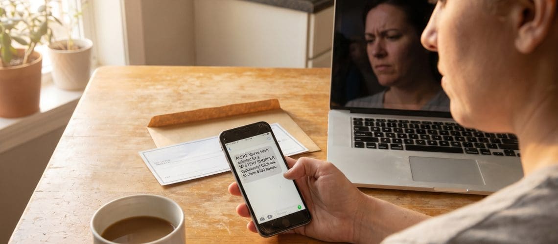 A mystery shopper at a kitchen table examining a suspicious check and torn envelope with a wary, skeptical expression — a laptop nearby showing an email. Conveys "spot the scam before you deposit.